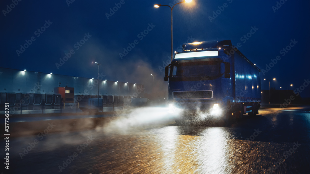 Blue Long Haul Semi-Truck with Cargo Trailer Full of Goods Travels At Night on the Freeway Road, Driving Across Continent Through Rain, Fog, Snow. Industrial Warehouses Area.