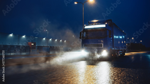 Blue Long Haul Semi-Truck with Cargo Trailer Full of Goods Travels At Night on the Freeway Road, Driving Across Continent Through Rain, Fog, Snow. Industrial Warehouses Area.