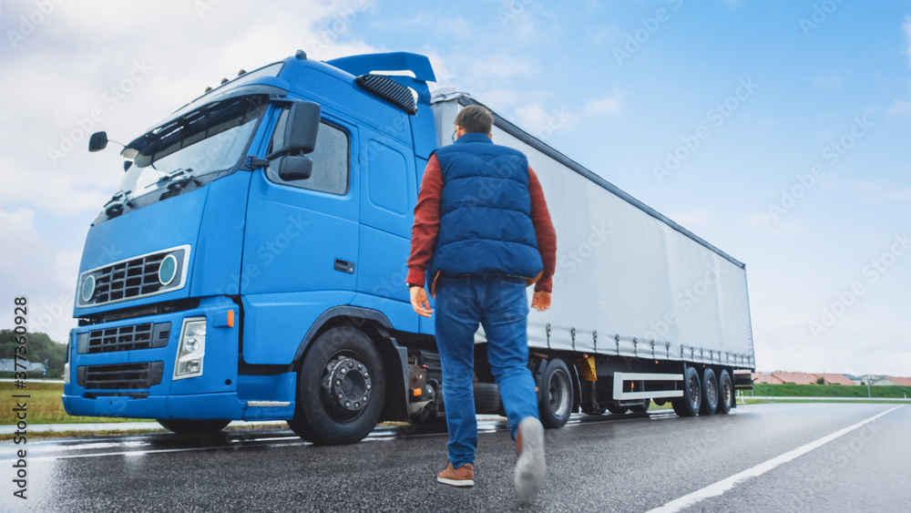 Truck Driver Crosses the Road in the Rural Area and Gets into His Blue Long Haul Semi-Truck with Cargo Trailer Attached. Logistics Company Moving Goods Across Countrie and Continent