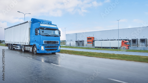 Long Haul Semi-Truck with Cargo Trailer Full of Goods Travels on the Highway Road. Daytime Driving Across Continent Through Rain. Industrial Retail Warehouses Area. Front Shot