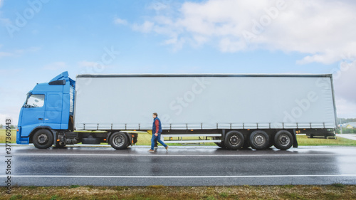 Truck Driver Crosses the Road in the Rural Area and Gets into His Blue Long Haul Semi-Truck with Cargo Trailer Attached. Logistics Company Moving Goods Across Countrie Continent. Side View Shot