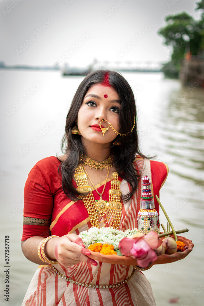 Portrait of beautiful Indian girl standing in front of ganga river wearing traditional Indian saree, gold jewellery and bangles holding plate of religious offering. Maa Durga agomoni shoot concept