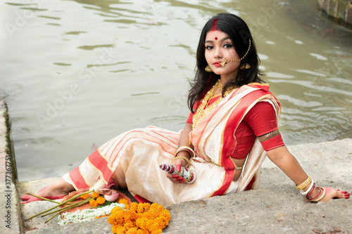 Portrait of beautiful Indian girl in front of ganga river wearing traditional Indian saree, gold jewellery and bangles holding plate of religious offering. Maa Durga agomoni shoot concept photo