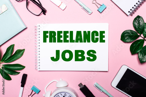 FREELANCE JOBS written in green on a pink background. Business attributes and green plants. Flat lay. Business concept