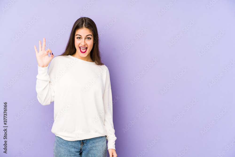 Young caucasian woman isolated on purple background winks an eye and holds an okay gesture with hand. Young caucasian woman isolated on purple background winks an eye and holds an okay gesture with hand.