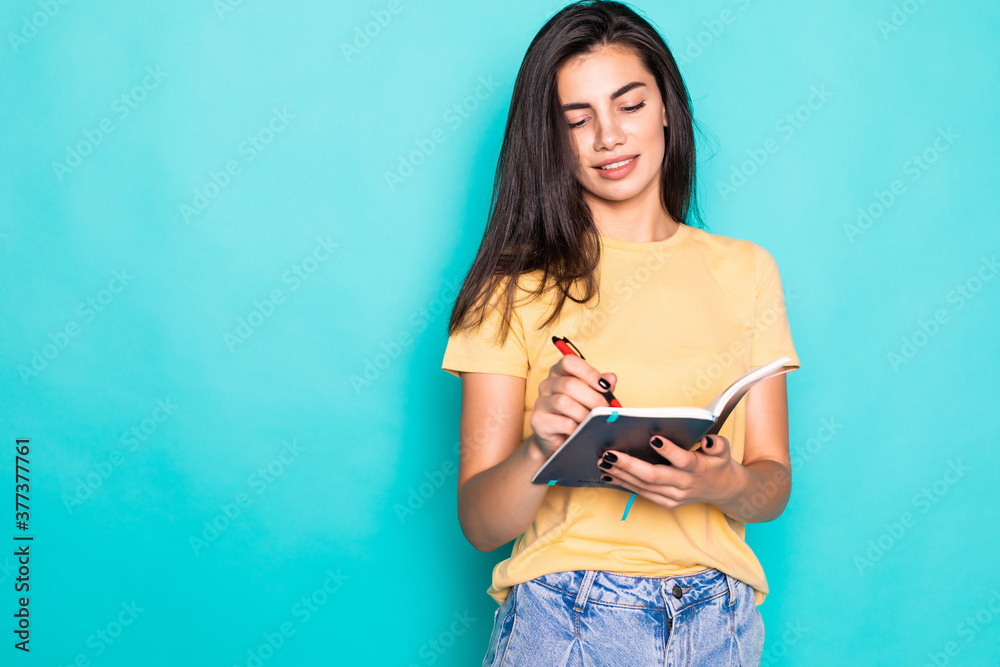 Pretty young beautiful woman standing, writing, take notes, holding textbook notebook organizer in hand and pen on turquoise background Pretty young beautiful woman standing, writing, take notes, holding textbook notebook organizer in hand and pen on turquoise background