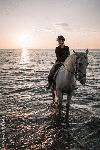 woman on a white horse stands in the sea at sunset