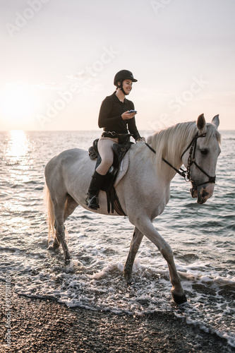 young woman with a phone in her hand rides astride a white beautiful horse and laughs on the beach at sunset
