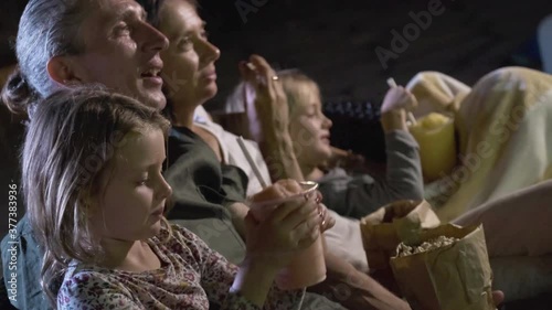 Family sitting on sofa enjoying movie night, eating popcorn and drinking milkshakes. Close up of mother, father and children watching projector TV or movies
