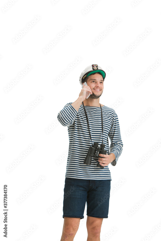 Fototapeta premium Talking phone. Caucasian male sailor in uniform on white studio background. Young man using modern devices and gadgets in his work. Concept of tech, professional occupation, job of seaman.