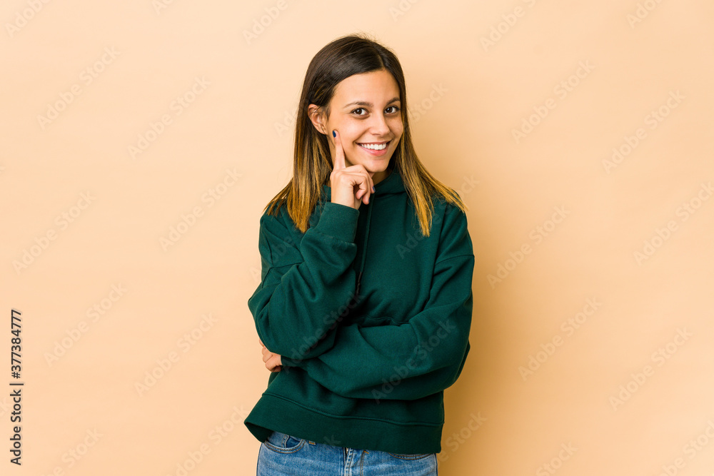 Young woman isolated on beige background smiling happy and confident, touching chin with hand.