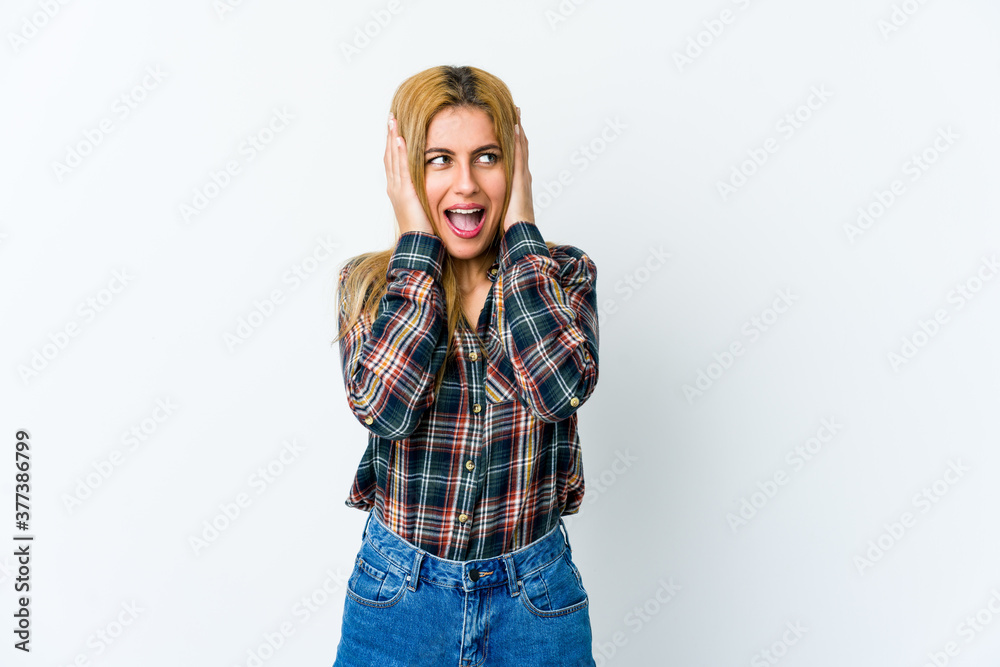 Young blonde woman isolated on white background covering ears with hands trying not to hear too loud sound.