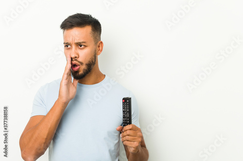Young south-asian man holding a tv controller is saying a secret hot braking news and looking aside