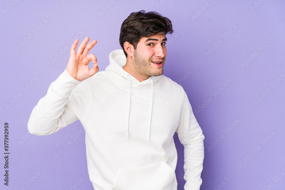 Young man isolated on purple background winks an eye and holds an okay gesture with hand. Young man isolated on purple background winks an eye and holds an okay gesture with hand.