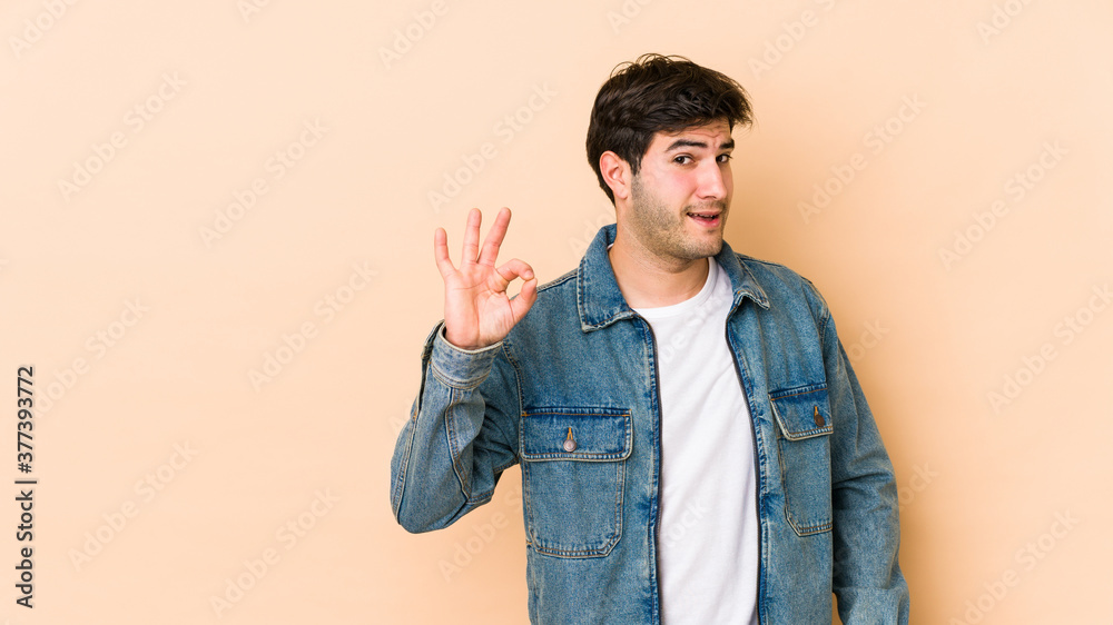 Young man isolated on beige background winks an eye and holds an okay gesture with hand. Young man isolated on beige background winks an eye and holds an okay gesture with hand.