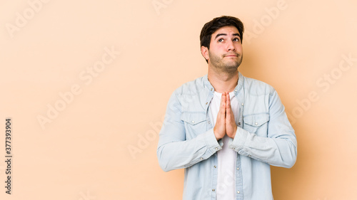 Young man isolated on beige background holding hands in pray near mouth, feels confident.