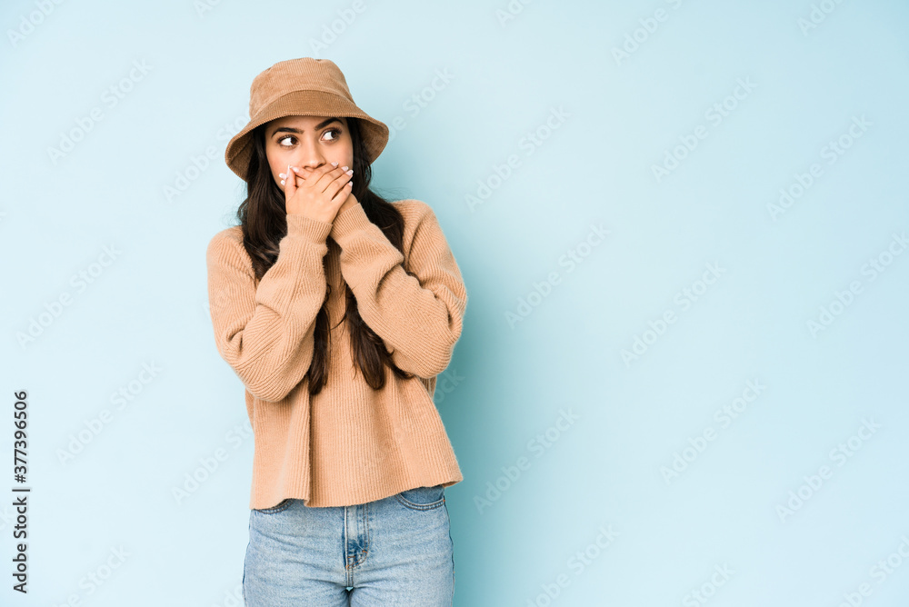 Young indian woman wearing a hat isolated on blue background thoughtful looking to a copy space covering mouth with hand. Young indian woman wearing a hat isolated on blue background thoughtful looking to a copy space covering mouth with hand.