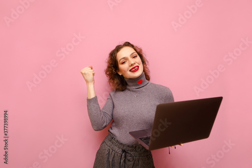 Joyful girl with curly red hair stands with a laptop in her hand6 looks into the camera and smiles. Positive lady freelancer rejoices with laptop in hand on pink background. Isolated. Copy space