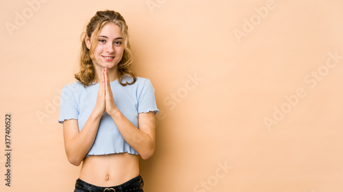 Young caucasian woman isolated on beige background holding hands in pray near mouth, feels confident.