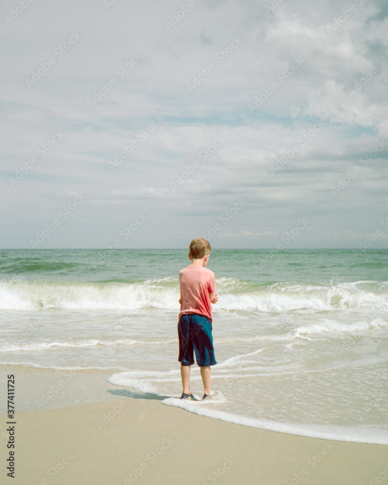 Boy standing on the beach looking trepidatiously at the water Boy standing on the beach looking trepidatiously at the water