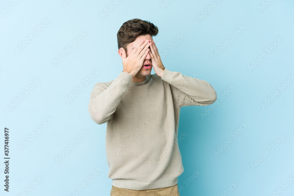 Young caucasian man isolated on blue background afraid covering eyes with hands.