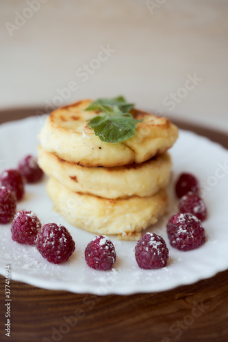 Juicy curd cheesecakes lie on a white saucer with raspberries and mint leaf. Light background