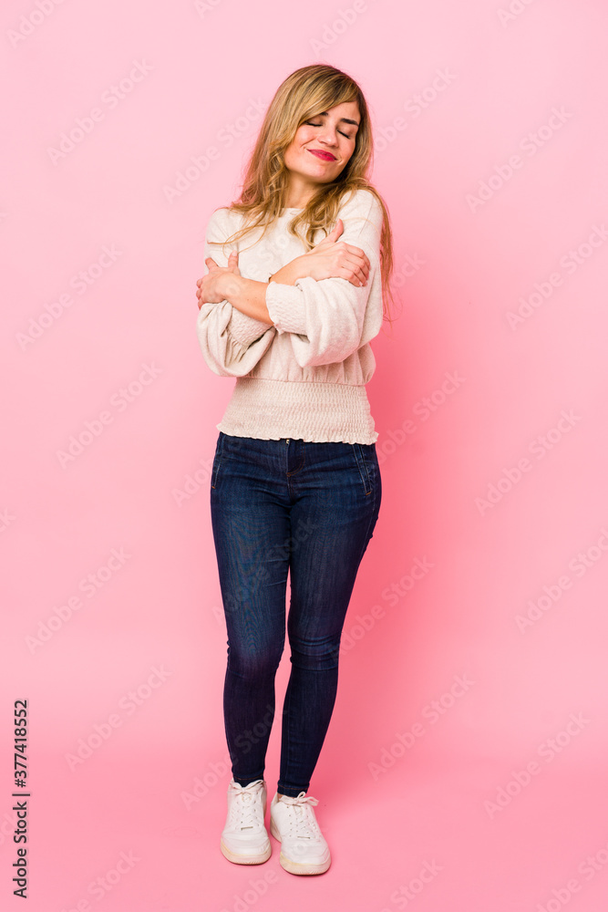 Young blonde caucasian woman standing over a pink background hugs, smiling carefree and happy.
