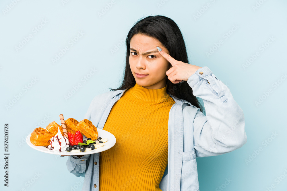 Young chinese woman holding waffle isolated on blue background pointing with finger at you as if inviting come closer. Young chinese woman holding waffle isolated on blue background pointing with finger at you as if inviting come closer.