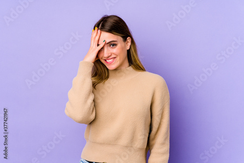 Young caucasian woman isolated on purple background blink at the camera through fingers, embarrassed covering face.