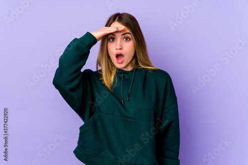 Young caucasian woman isolated on purple background looking far away keeping hand on forehead. © Asier