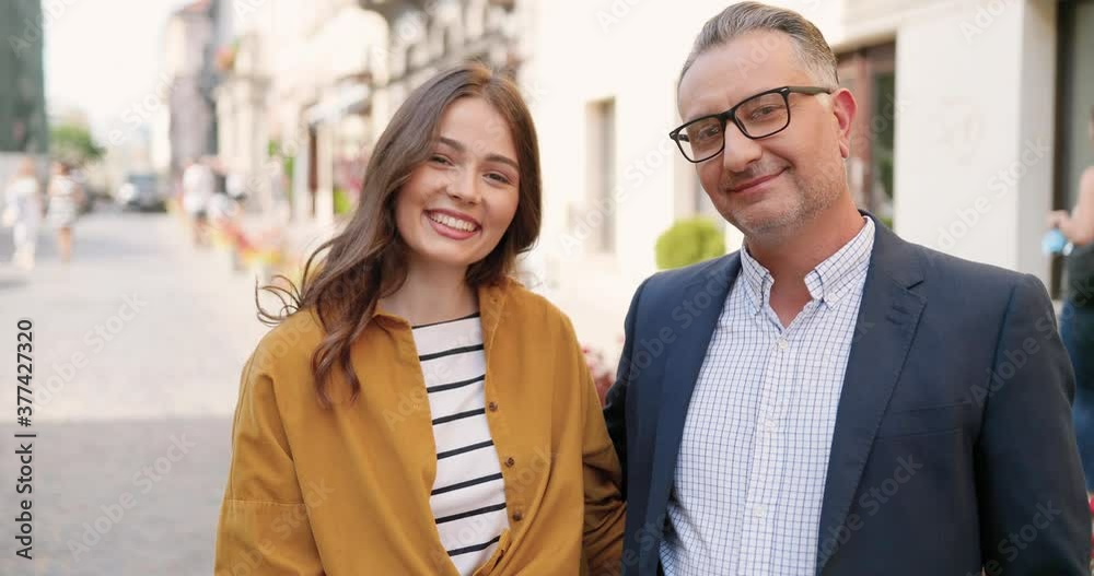 Portrait of Caucasian handsome middle-aged father in glasses standing at city street and hugging pretty young adult daughter. Outdoors. Man and girl with smiles. Fatherhood love concept. Portrait of Caucasian handsome middle-aged father in glasses standing at city street and hugging pretty young adult daughter. Outdoors. Man and girl with smiles. Fatherhood love concept.