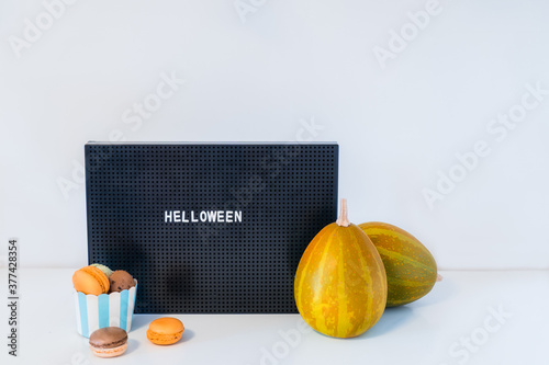 Minimalist Halloween decor - letter board with Helloween message, decorative pumpkins and cup with sweet macaroons on white background. Festive concept. Copy space.