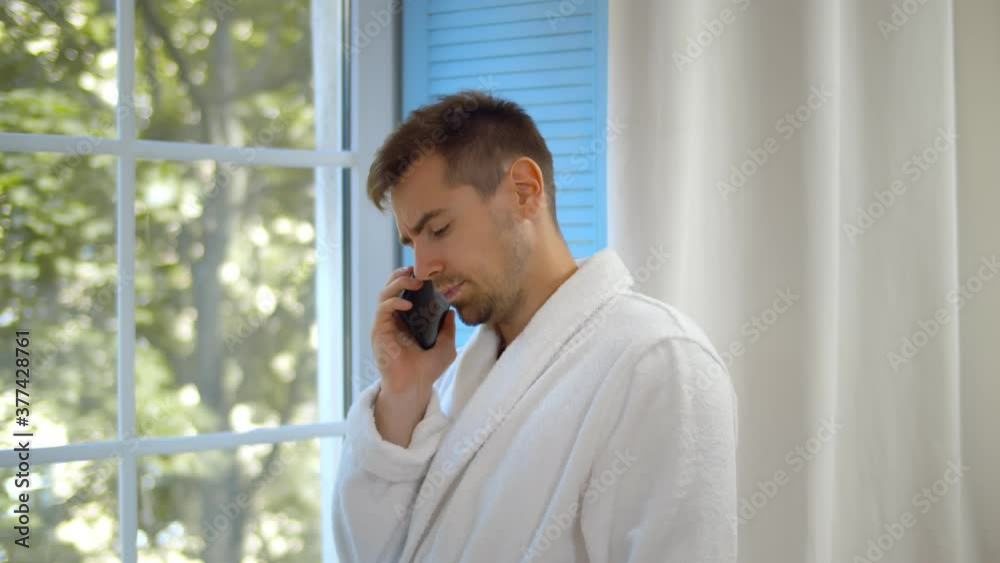 Smiling young attractive man wearing bathrobe talking on mobile phone while standing at window Smiling young attractive man wearing bathrobe talking on mobile phone while standing at window