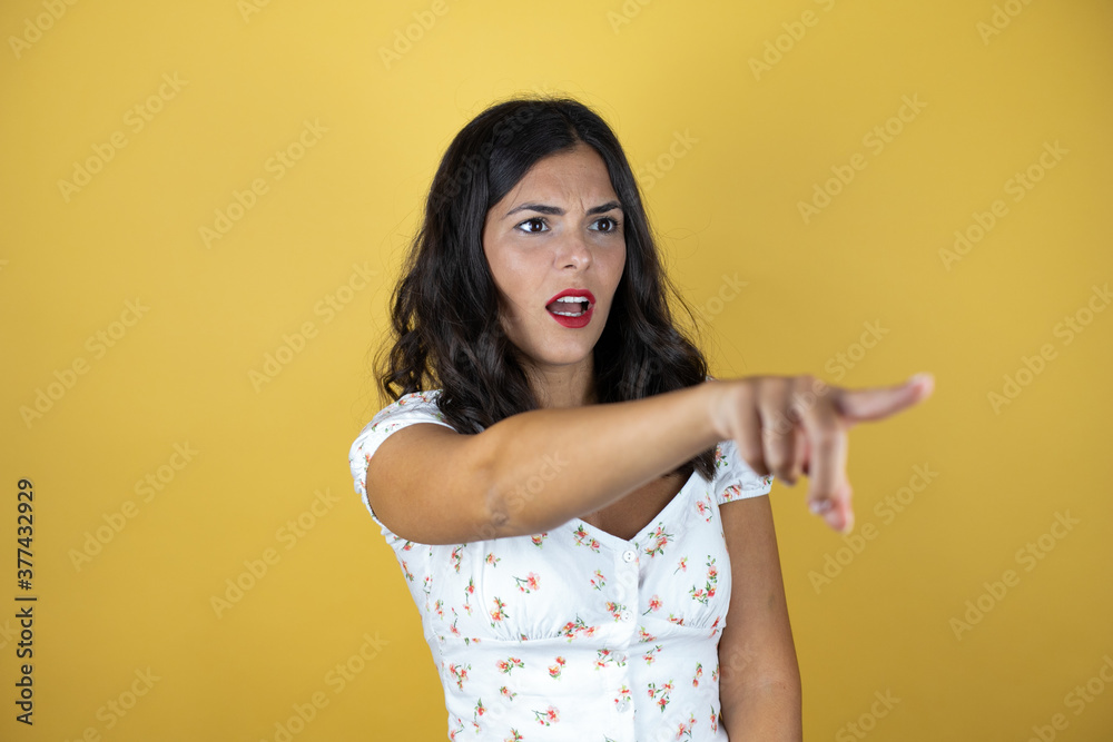 Beautiful woman over yellow background pointing with finger surprised ahead, open mouth amazed expression, something on the front