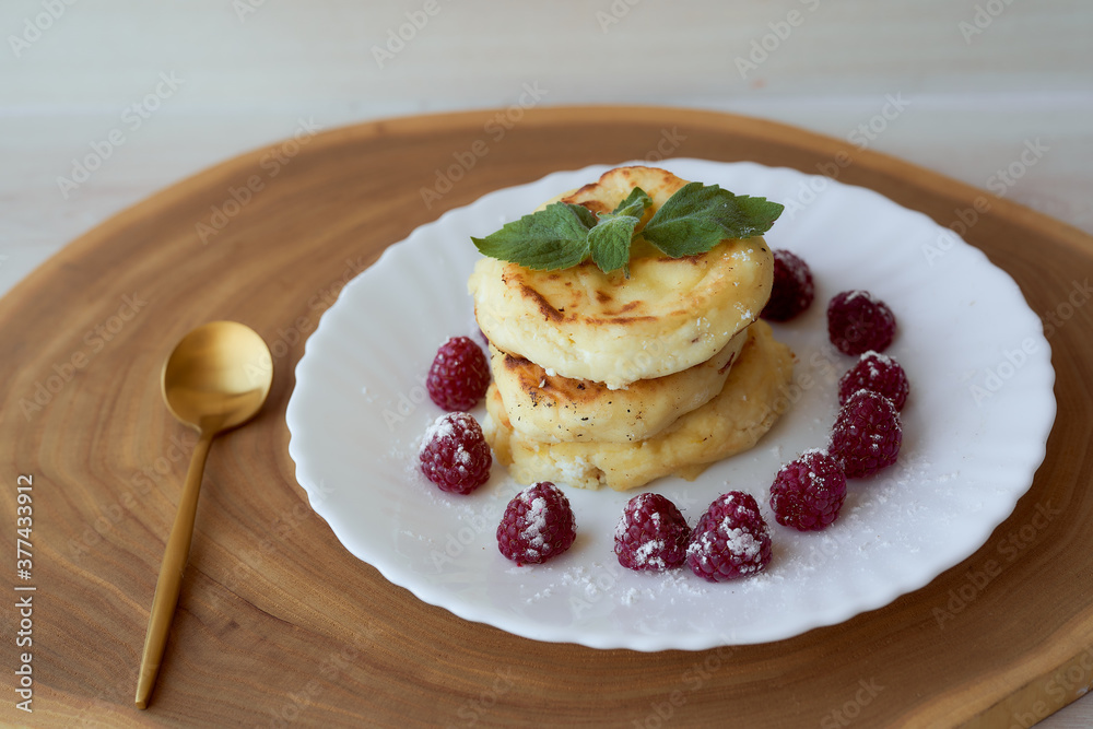 Juicy curd cheesecakes lie on a white saucer with raspberries and mint leaf. With golden spoon. Light background Juicy curd cheesecakes lie on a white saucer with raspberries and mint leaf. With golden spoon. Light background