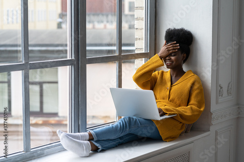 Happy African American woman freelancer with afro hairstyle wear yellow cardigan sitting on windowsill, working on laptop, talking in video chat, laughing with toothy smile. Forgot, bad memory concept photo