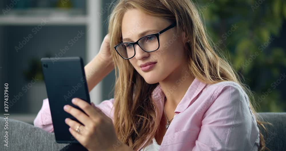 Attractive lovely girl with blond hair using digital tablet while sitting on cozy sofa. Happy woman in casual clothing and eyewear spending free time online. Attractive lovely girl with blond hair using digital tablet while sitting on cozy sofa. Happy woman in casual clothing and eyewear spending free time online.