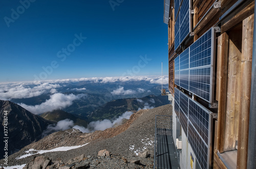 Fototapeta Solar panels mounted on the mountain refuge hut walls providing necessary electricity for a guest' needs with wide-angle Alps mountains landscape view