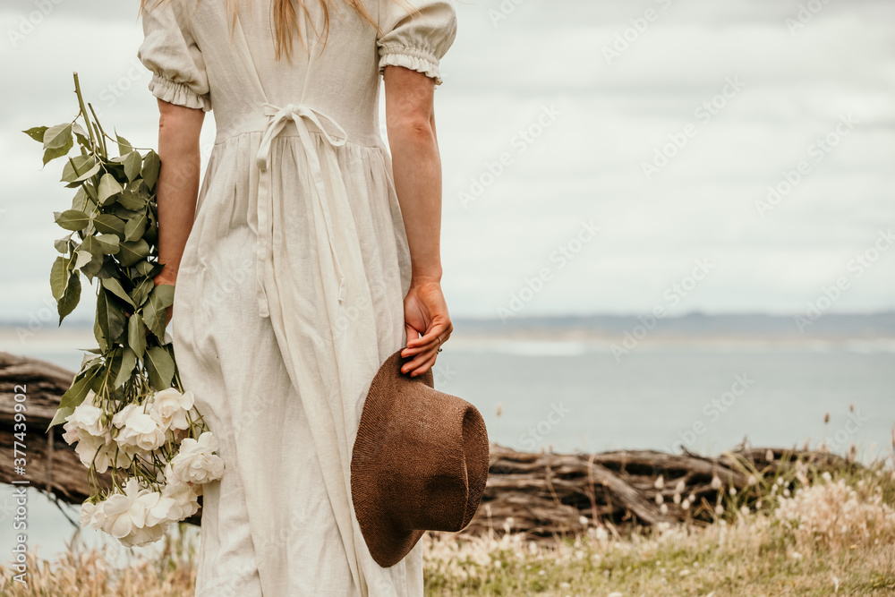 Lady holds flowers and a hat by the sea.