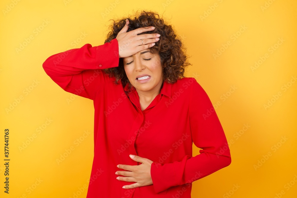 Young arab woman with curly hair wearing red shirt on yellow background Touching forehead for illness and fever, flu and cold, virus sick. Young arab woman with curly hair wearing red shirt on yellow background Touching forehead for illness and fever, flu and cold, virus sick.