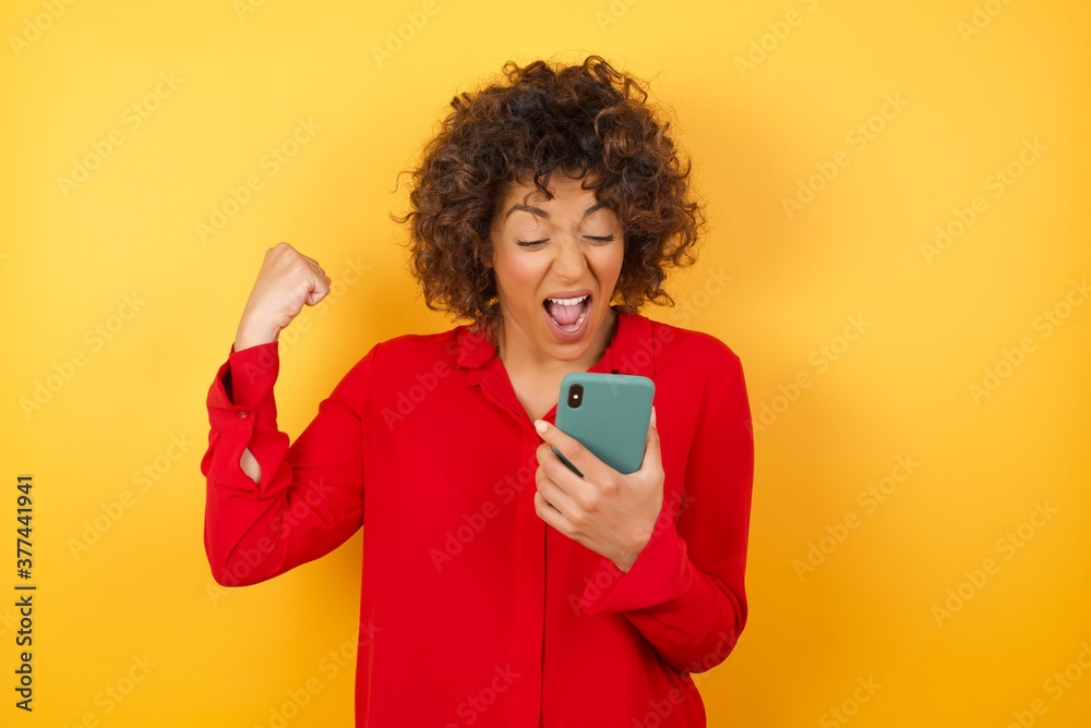 Portrait of Young arab woman with curly hair wearing red shirt on yellow background holding in hands cell reading browsing Portrait of Young arab woman with curly hair wearing red shirt on yellow background holding in hands cell reading browsing
