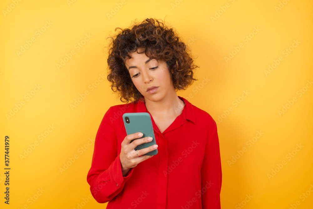 Portrait of excited Young arab woman with curly hair wearing red shirt on yellow background winking and eye hold smart phone use read social network news Portrait of excited Young arab woman with curly hair wearing red shirt on yellow background winking and eye hold smart phone use read social network news