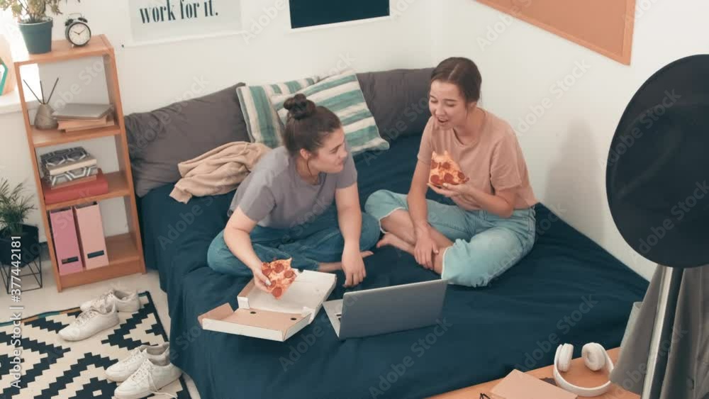 High angle shot of happy young women sitting cross-legged on bed in cozy bedroom and laughing while eating pizza and watching something funny on laptop