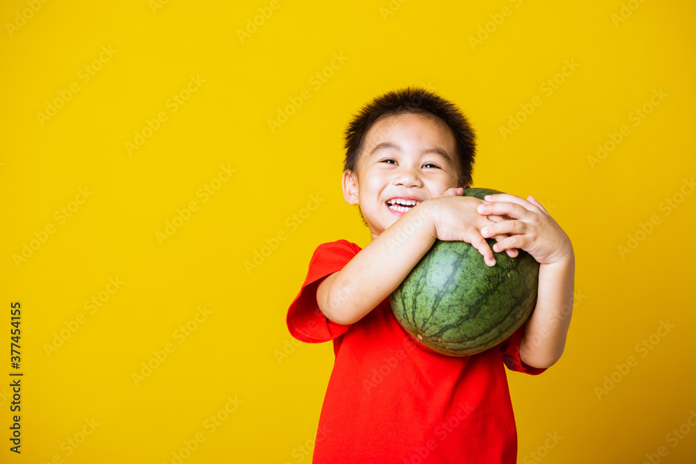 Happy portrait Asian child or kid cute little boy attractive smile wearing red t-shirt playing holds full watermelon that has not been cut, studio shot isolated on yellow background Happy portrait Asian child or kid cute little boy attractive smile wearing red t-shirt playing holds full watermelon that has not been cut, studio shot isolated on yellow background