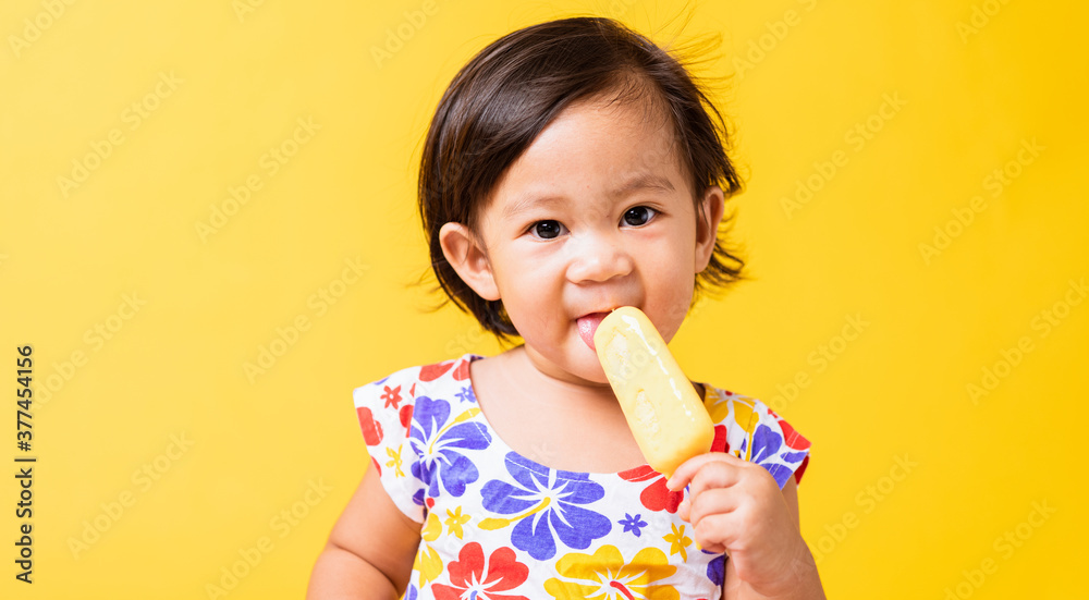 Happy portrait Asian baby or kid cute little girl attractive laugh smile wearing dick pattern shirt holds and eating sweet wooden ice cream, studio shot isolated on yellow background, summer concept Happy portrait Asian baby or kid cute little girl attractive laugh smile wearing dick pattern shirt holds and eating sweet wooden ice cream, studio shot isolated on yellow background, summer concept