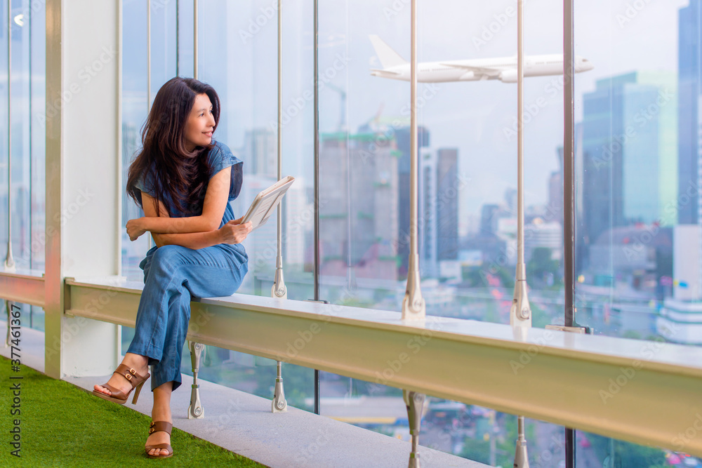 Happy asian woman holding tablet computer and looking away at copy space while sitting near window glass with airplane and business buildings on background.