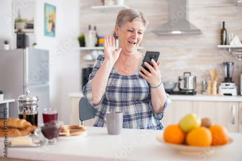 Senior woman waving during video conference using smartphone in kitchen while having breaksfat. Elderly person using internet online chat technology video webcam making a video call connection camera