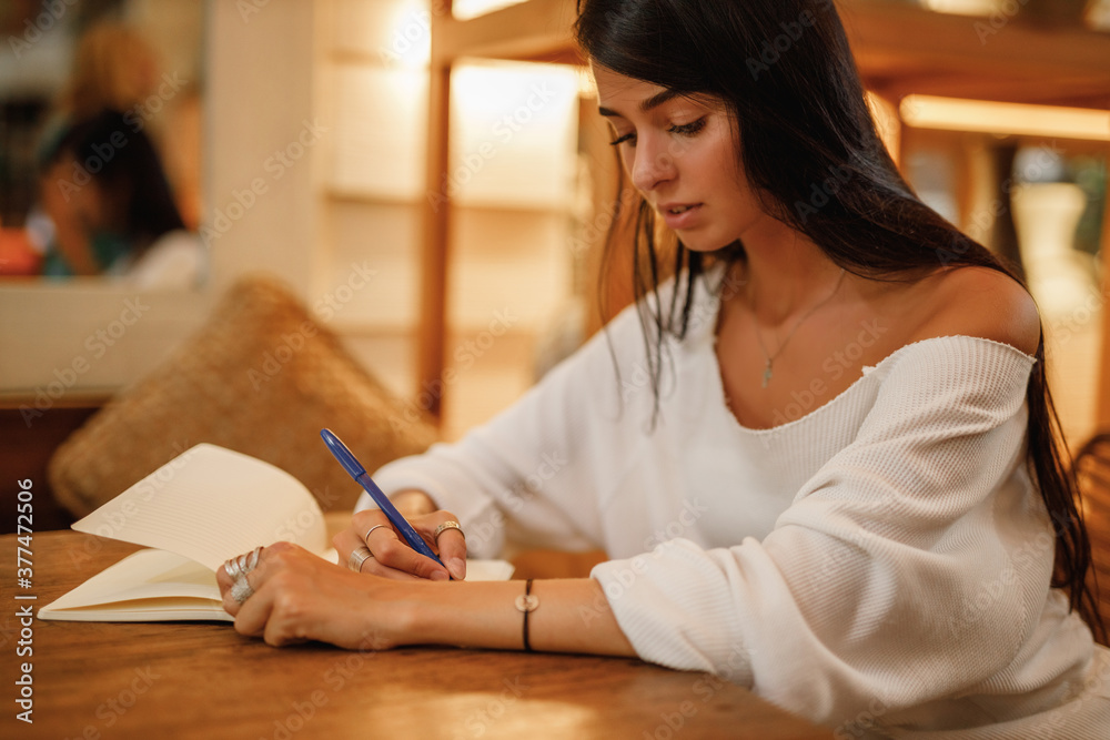 Positive female student writing down story in notebook sitting in coffee shop . Girl noting text in notepad Positive female student writing down story in notebook sitting in coffee shop . Girl noting text in notepad