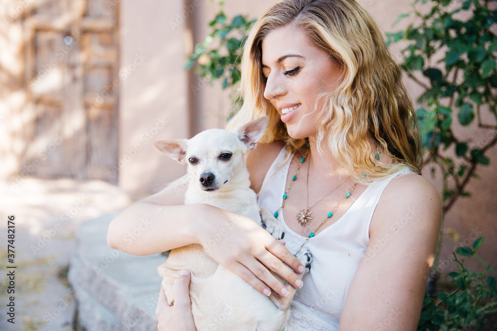 Young blond haired woman holding cuddling a blond short haired Chihuahua dog in her arms with loving affection. She is outdoors in a southwestern beige and light green environment. She is wearing an Young blond haired woman holding cuddling a blond short haired Chihuahua dog in her arms with loving affection. She is outdoors in a southwestern beige and light green environment. She is wearing an
