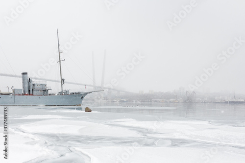 The ship stands on the dock, the sea is covered with ice, it snows, the far bank in the fog